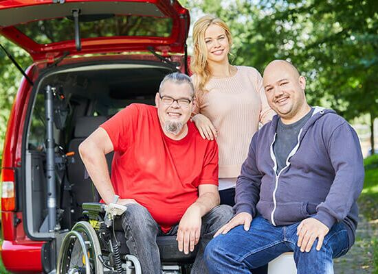 Ein Klient im Rollstuhl zusammen mit zwei Kolleg:innen vom Fahrdienst stehen an der offenen Caddy-Hackklappe