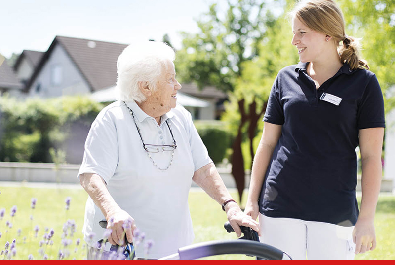 Eine Betheljahr-Teilnehmerin begleitet eine ltere Dame am Rollator im Garten einer Senioren-Einrichtung